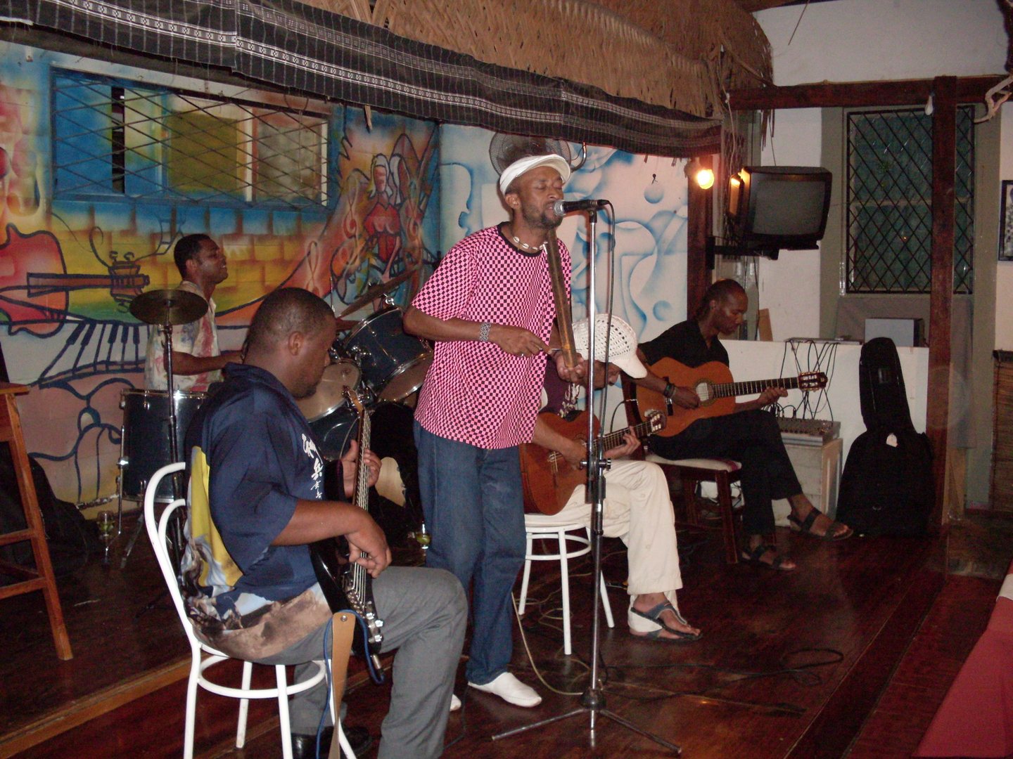 Bino Branco singing into a microphone and holding a metal scraper, with seated string players and a drummer