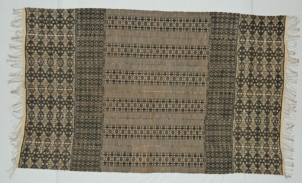 A rectangular Cabo Verde panu d’obra assembled from six narrow woven strips, patterned in black and cream with diamonds, chevrons, and fringed ends